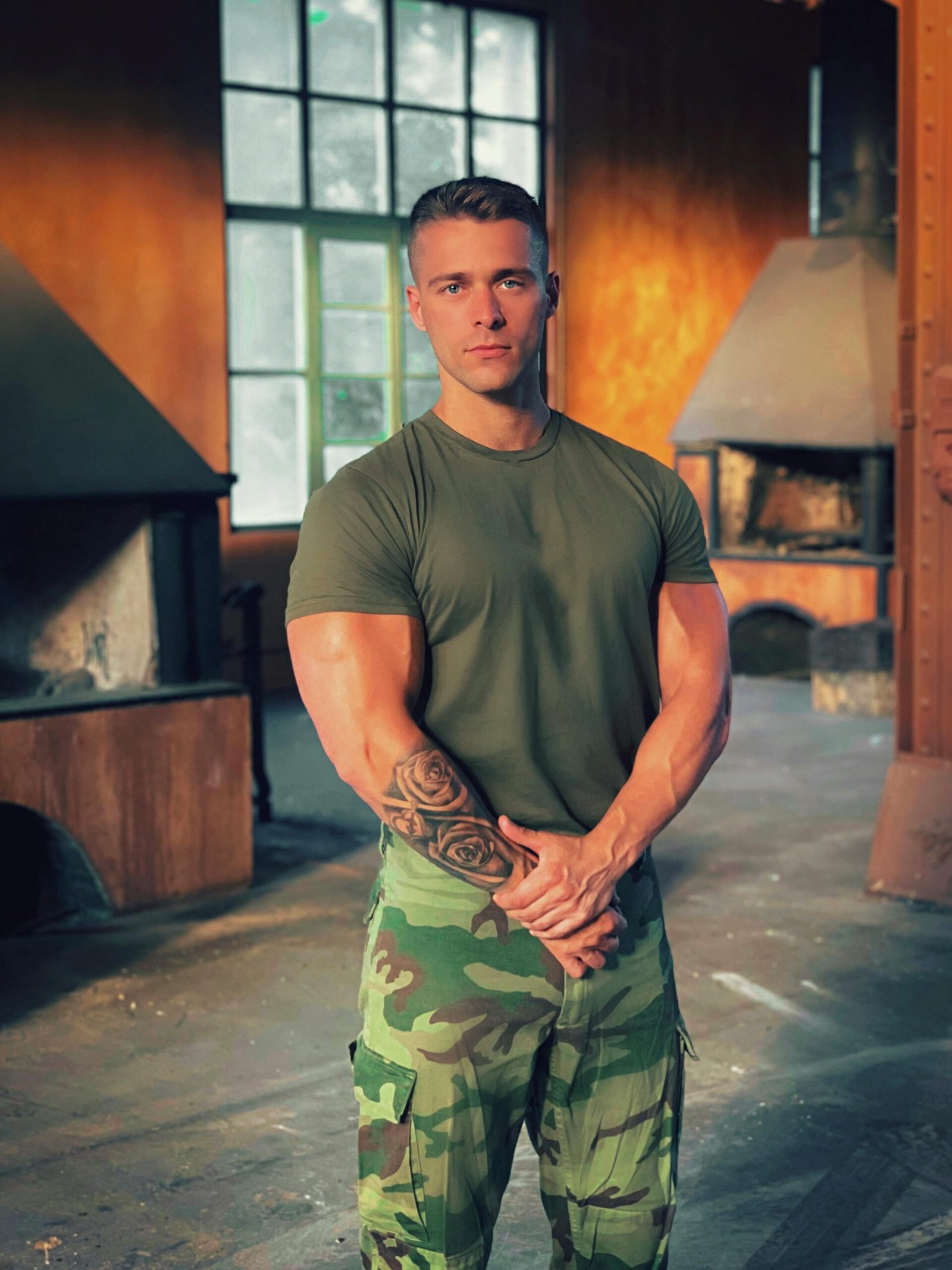 A muscular man in camo pants confidently poses indoors, showcasing his tattoos and strong build.